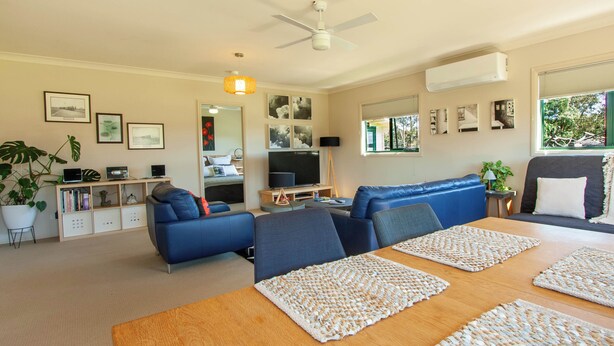 Ein modernes Wohnzimmer mit einem blauen Sofa, einem Holzesstisch mit gewebten Tischsets, einem Fernseher und einem Deckenventilator.