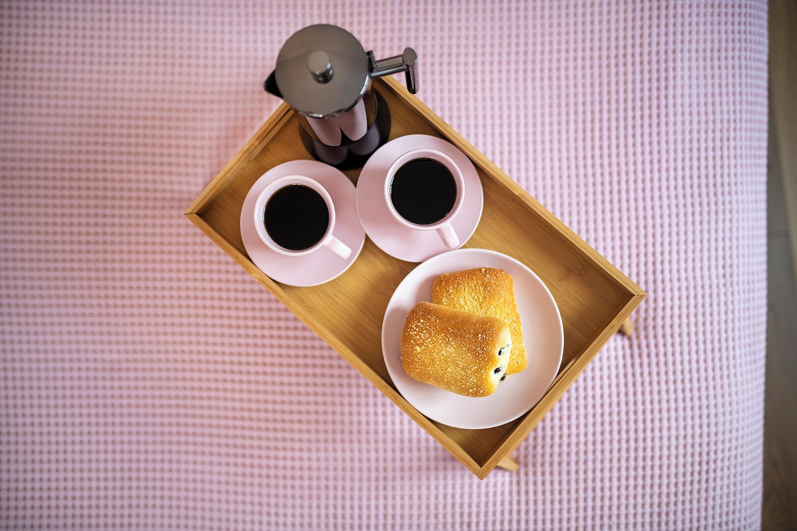 A wooden tray with two cups of coffee and a pastry on a pink textured surface.