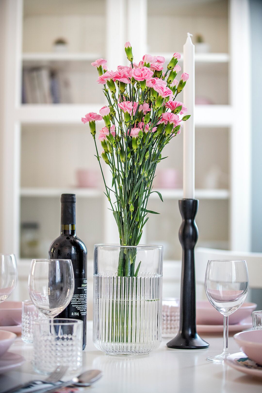 A table with a clear glass vase containing pink flowers, a candle, and a bottle of wine.