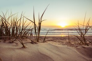 A beach with sand and grass in the foreground and a sunset over the ocean in the background. - Cool Waves: A fab contemporary holiday home in Rhosneigr (Rhosneigr)