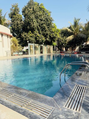 A swimming pool with clear blue water, surrounded by trees and a building with a tiled exterior. - Nilkanth Resort - Best Resort in Anand (Anand)