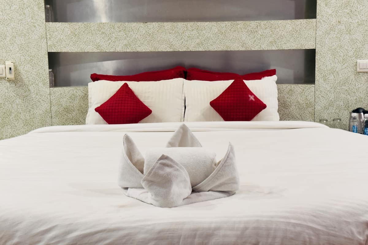 Ein ordentlich bezogenes Bett mit weißer Decke und zwei roten Kissen, ein graues Handtuch sowie ein moderner Kopfseitenverkleiderung mit integriertem Ablagefach.