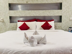 Ein ordentlich bezogenes Bett mit weißer Decke und zwei roten Kissen, ein graues Handtuch sowie ein moderner Kopfseitenverkleiderung mit integriertem Ablagefach.