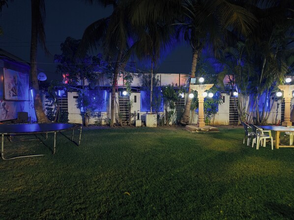 An outdoor area with a grassy lawn, a table with chairs, and a stage with a backdrop. - Nilkanth Resort - Best Resort in Anand (Anand)