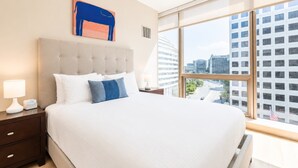 A hotel room with a large bed, a nightstand with a lamp, a digital clock, and a city view through large windows. - Luxury Apt Near Downtown (Stamford, CT)