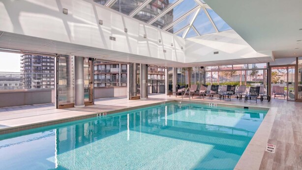An indoor swimming pool with clear glass ceilings, lounge chairs, and a view of surrounding buildings. - Luxury Apt Near Downtown (Stamford, CT)