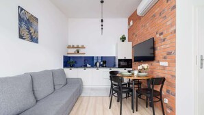A compact living space with a grey sofa, a dining table with four chairs, a kitchen area with a TV, and a brick accent wall.