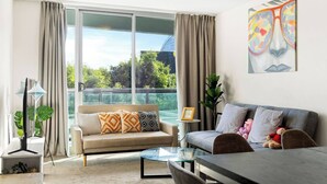 Una sala de estar moderna con un sofá, una mesa de comedor y una mesa de centro de vidrio. Hay una ventana grande con cortinas, una planta y un cuadro en la pared.