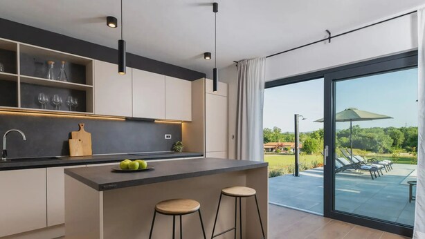 Eine moderne Küche mit einer zentralen Kochinsel, Barhocker und Blick auf einen Garten durch große Schiebeglasüren.