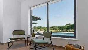 Ein Zimmer mit zwei grünen Stühlen, einem kleinen Tisch mit einer Flasche und einer Tasse, und Blick auf ein Schwimmbad und Grünflächen durch große Fenster.