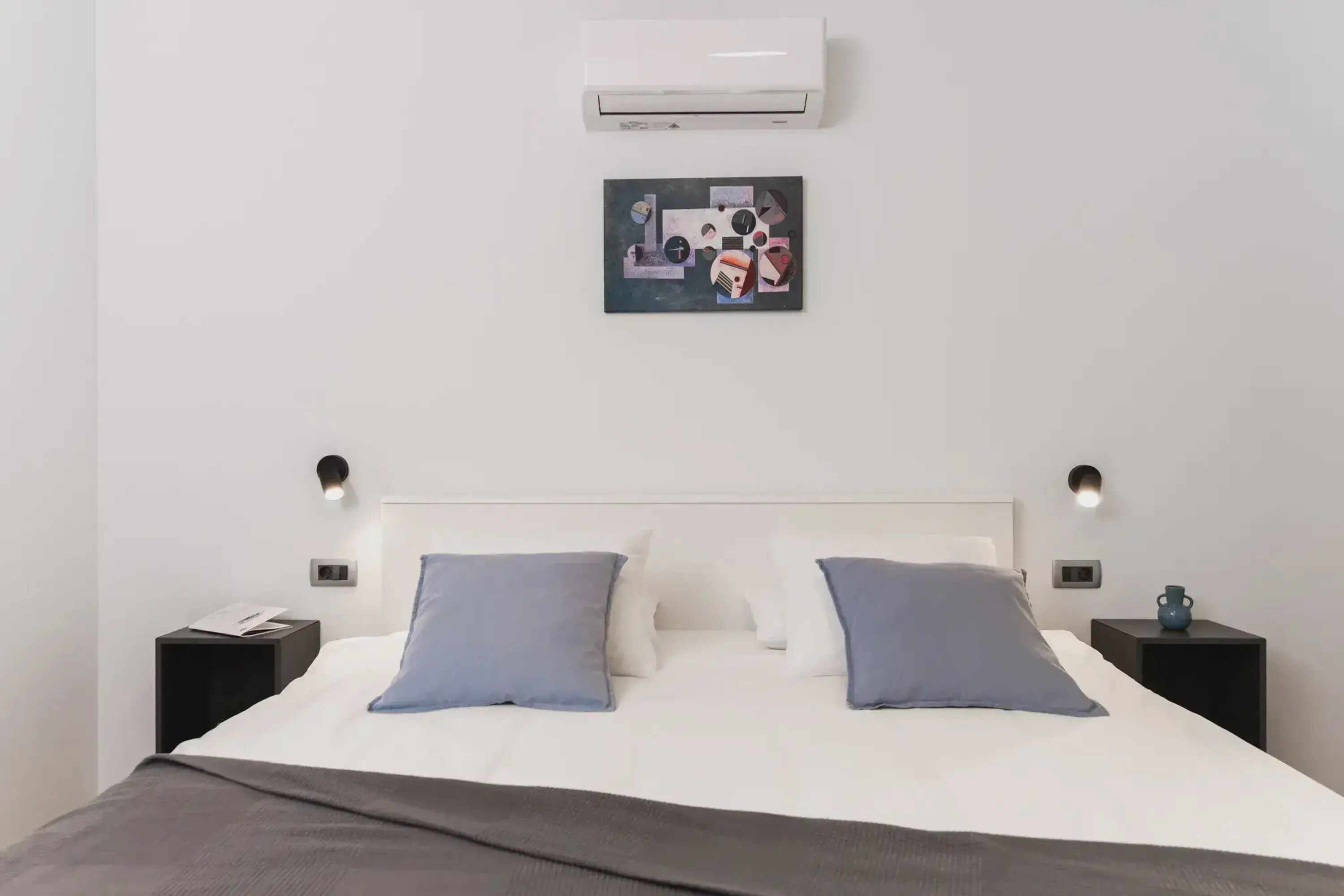 A modern hotel room with a white bed, two pillows, a gray blanket, a wall-mounted air conditioner, a small painting, and two bedside tables with lamps.