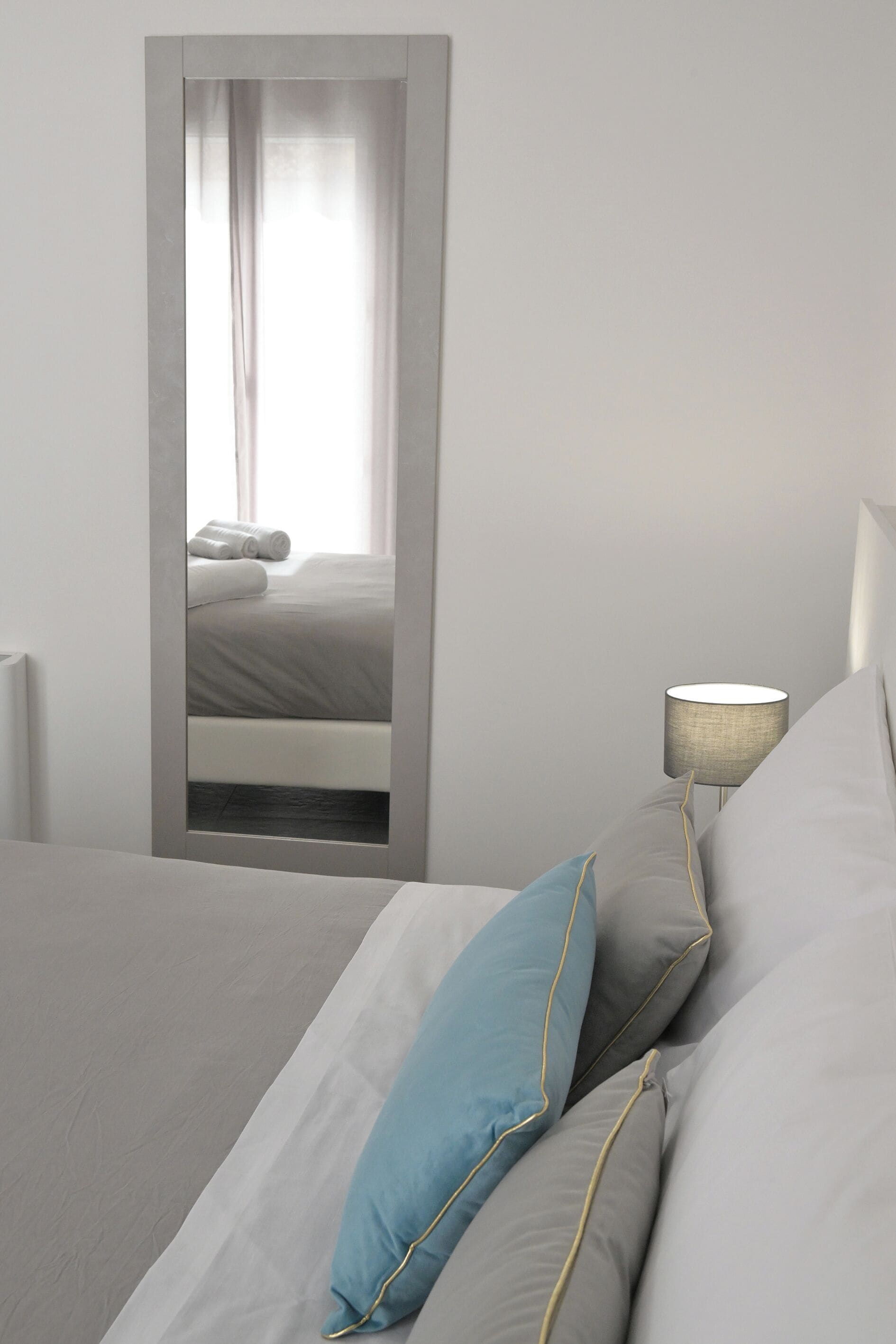 Een net opgemaakt bed met kussens, een bedlampje en een spiegel die een raam met gordijnen reflecteert.
