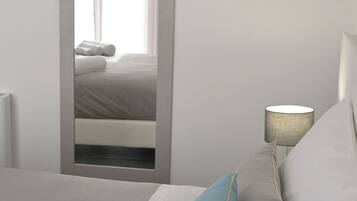 Een net opgemaakt bed met kussens, een bedlampje en een spiegel die een raam met gordijnen reflecteert.