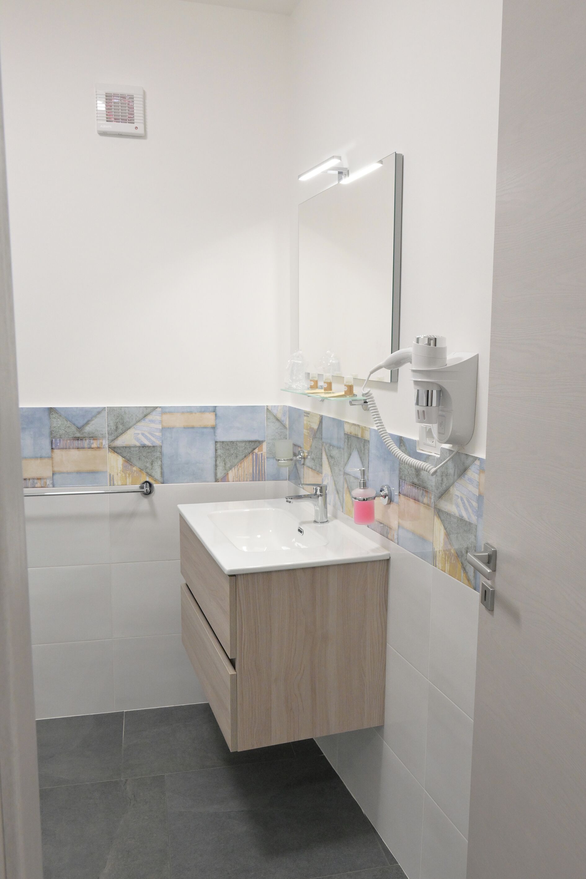 Un baño con lavamanos blanco, tocador de madera, salpicadero de azulejos y espejo con iluminación integrada.