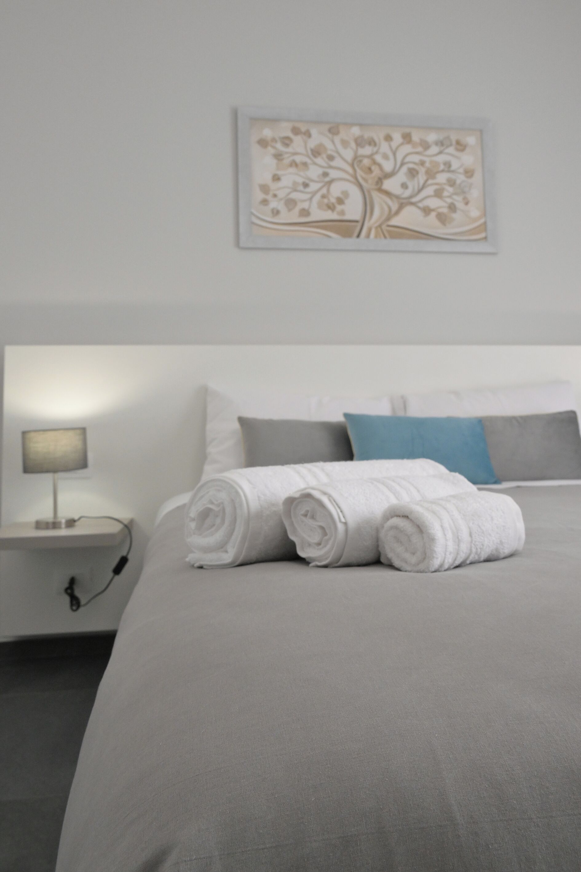 Una cama bien hecha con toallas blancas enrolladas, un cabecero gris y una almohada azul.