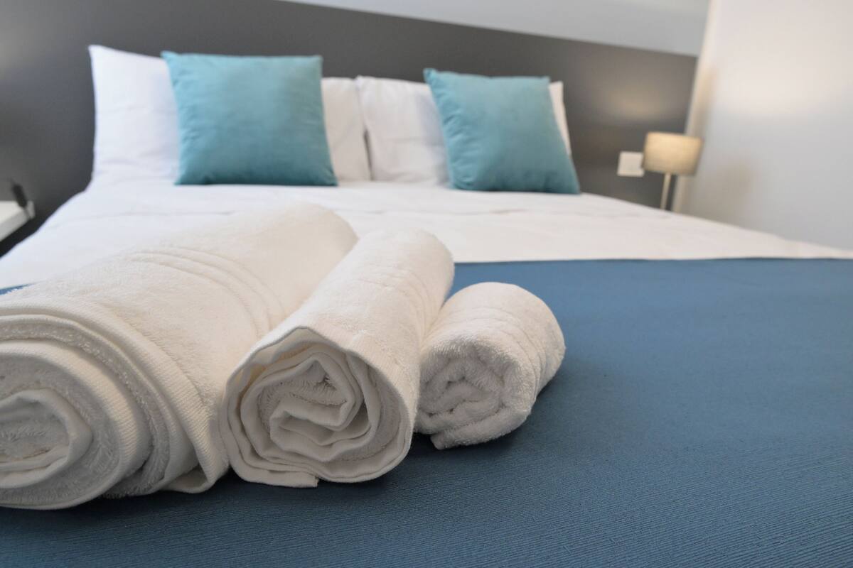 A neatly made bed with white and blue bedding, a blue bedspread, and a headboard with multiple pillows.
