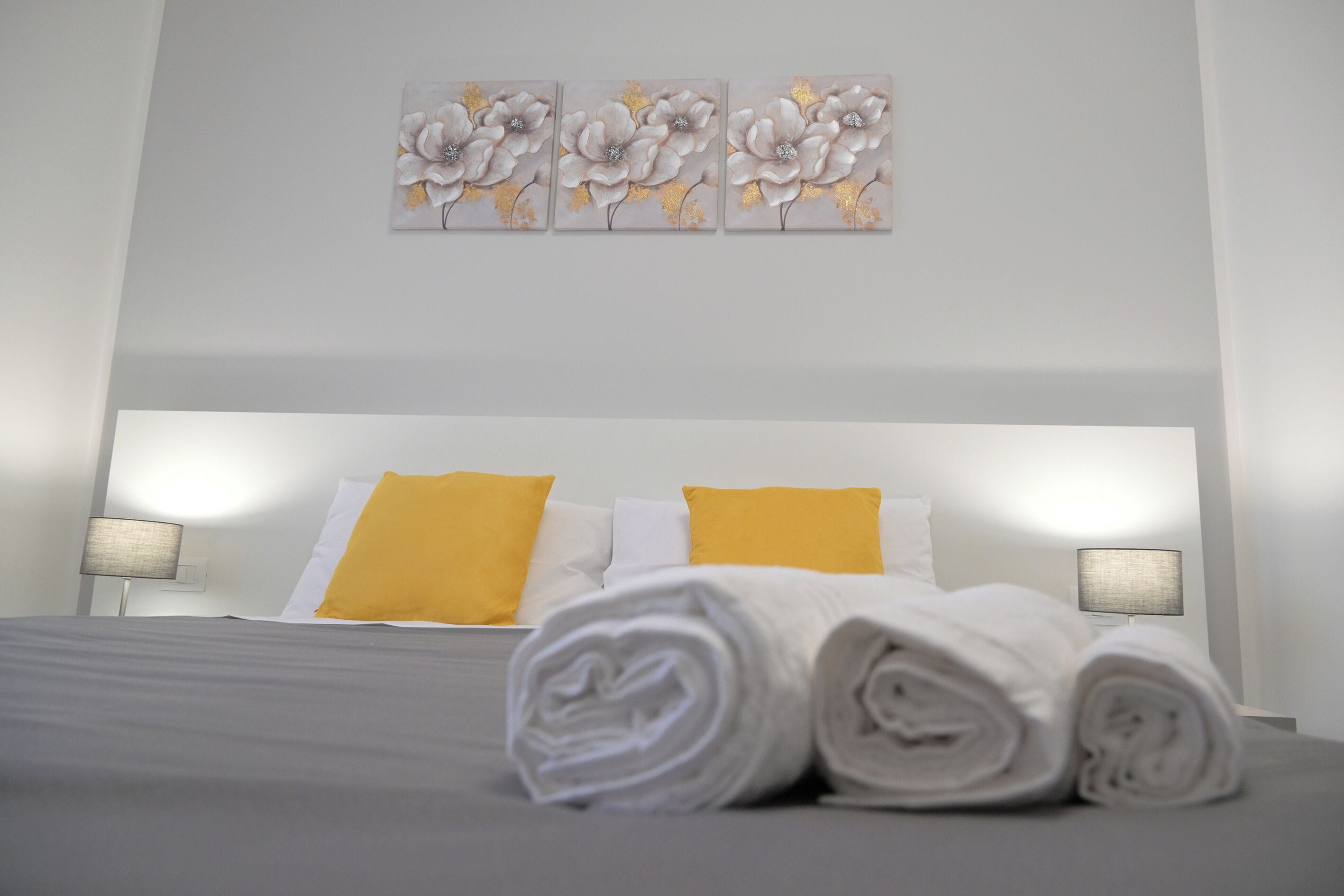 A neatly made bed with white and yellow pillows, a grey bedspread, and rolled white towels in front. There are three wall-mounted paintings above the bed.