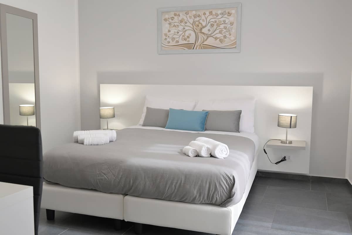 A neatly made bed with a grey and white color scheme, two bedside lamps, and a wall-mounted artwork of a tree.