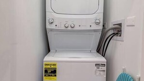 A compact laundry room with a front-loading washer and dryer, a blue ironing board, and a small shelf with a box on top. - Suite 2 on Southport 4 Queen Beds (Chicago)