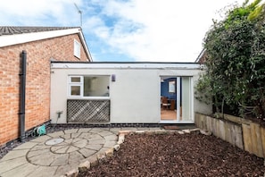 A small, single-story house with a brick exterior, a white extension with a window, and a garden area with mulch. - Inviting and Cosy Annexe | Free Parking and Wi-Fi (Nottingham)