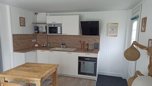 Een kleine keuken met witte kasten, een magnetron, een fornuis en een vaatwasser. Er staat een houten eettafel met twee stoelen.