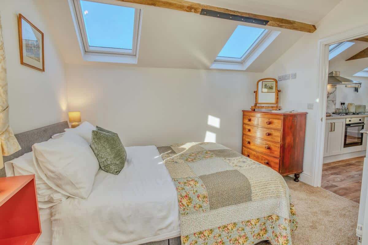 A bedroom with a skylight, a bed with a floral quilt, a wooden dresser, and a view into the kitchen.