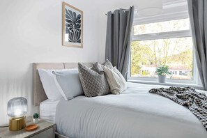 A neatly made bed with patterned pillows, a bedside lamp, and a window with curtains. - Spacious and Bright Home | Perfect for Contractors (Nottingham)