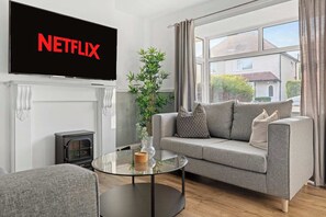 A living room with a sofa, a glass coffee table, a fireplace, a TV mounted on the wall, and a large window. - Spacious and Bright Home | Perfect for Contractors (Nottingham)