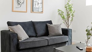 Een moderne woonkamer met een grijze bank, twee ingelijste kunstwerken, een kleine plant en een afstandsbediening op een bijzettafeltje.