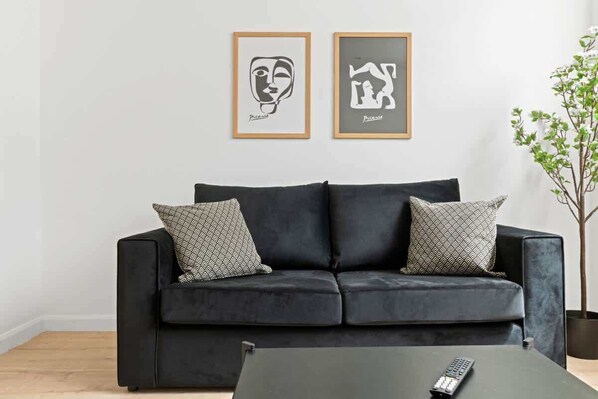 A black sofa with patterned pillows, a remote control on a coffee table, and two framed artworks on the wall.