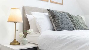 Een net opgemaakt bed met witte lakens, een sierkussen met een patroon, een bedlampje en een vaas met bloemen op een nachtkastje.