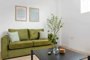 A green sofa with patterned pillows, a black coffee table with a remote control, a vase with a plant, and two framed artworks on the wall.