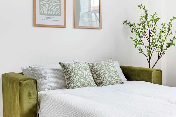 A bed with a white quilt, two patterned pillows, a green armchair, and a wall-mounted plant.