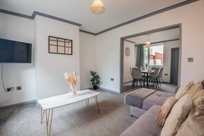 Una sala de estar con sofá, un comedor con sillas, un televisor montado en la pared y una mesa con un florero.