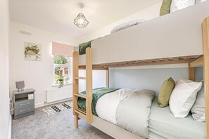 Een stapelbed met houten frame, een nachtkastje met lamp, een radiator en een raam met uitzicht op groen.