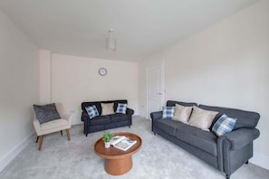 A living room with a beige armchair, two dark sofas with patterned cushions, a wooden coffee table with a magazine and a plant, and a white door.