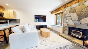 A modern living room with a stone fireplace, a sofa, a coffee table, and a kitchen area with wooden cabinets and a microwave. - Updated 2-Story Home, Great Views, Steam Shower & Washer/Dryer, Near Village (Mammoth Lakes)