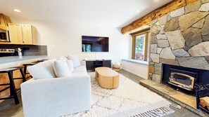A modern living room with a stone fireplace, a sofa, a coffee table, and a kitchen area with wooden cabinets and a microwave. - Updated 2-Story Home, Great Views, Steam Shower & Washer/Dryer, Near Village (Mammoth Lakes)