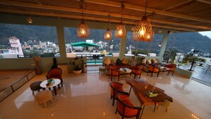 Un vaste espace extérieur aménagé avec de nombreuses tables et chaises, de grandes fenêtres offrant une vue sur une ville nichée dans les montagnes, et des lampes en osier suspendues.