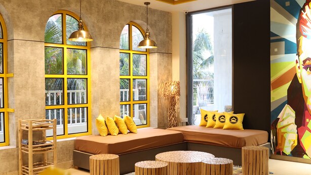 Un salon moderne avec un canapé d’angle, des tabourets en bois ronds et une grande fenêtre aux montures jaunes.