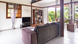 Een woonkamer met een grijze bank, een televisie, een boekenkast en een groot raam met uitzicht op groen.