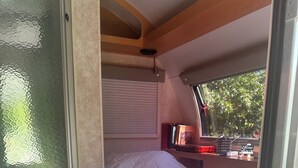 A compact RV bedroom with a bed, a small shelf with books, and a window with a view of trees. - Cozy RV on 149 acres in Charleston with AC, WiFi, and eight miles of trails!


 (Charleston)