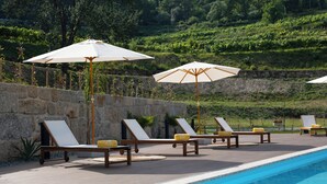 A poolside area with lounge chairs and umbrellas, a stone wall, and a view of a vineyard in the background. - Quinta de Reis (Ponte da Barca)