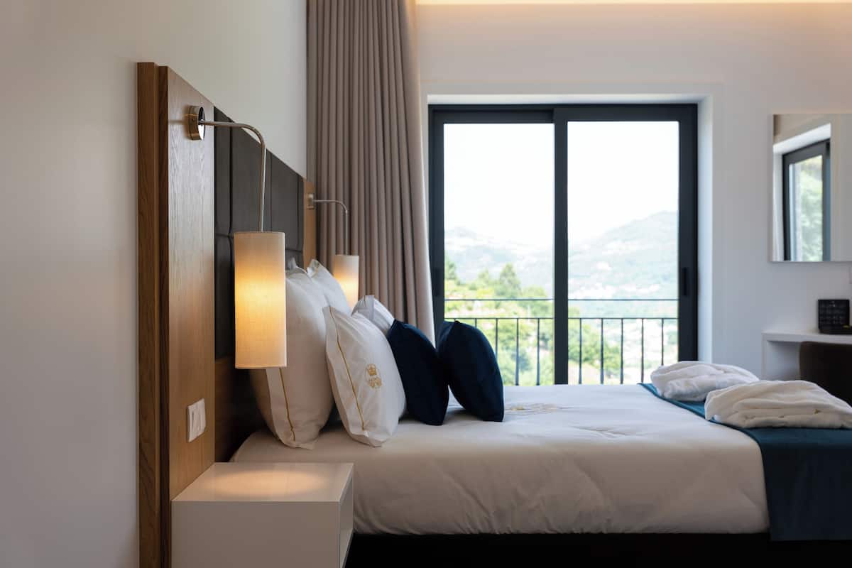 A hotel room with a bed, a nightstand, a lamp, a window with a view of the outdoors, and a mirror on the wall.
