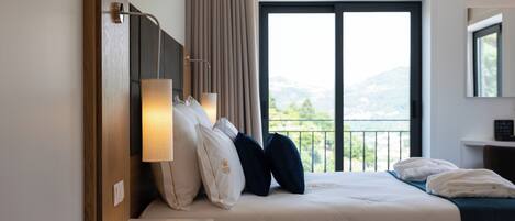 A hotel room with a bed, a nightstand, a lamp, a window with a view of the outdoors, and a mirror on the wall.