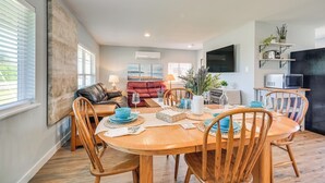 A modern living room with a dining area, featuring a wooden dining table, chairs, and a red sofa. There is a flat-screen TV mounted on the wall, and a kitchen area with a microwave and shelves. - Pet-friendly Moclips Home: Walk to Mocrocks Beach! (Moclips)
