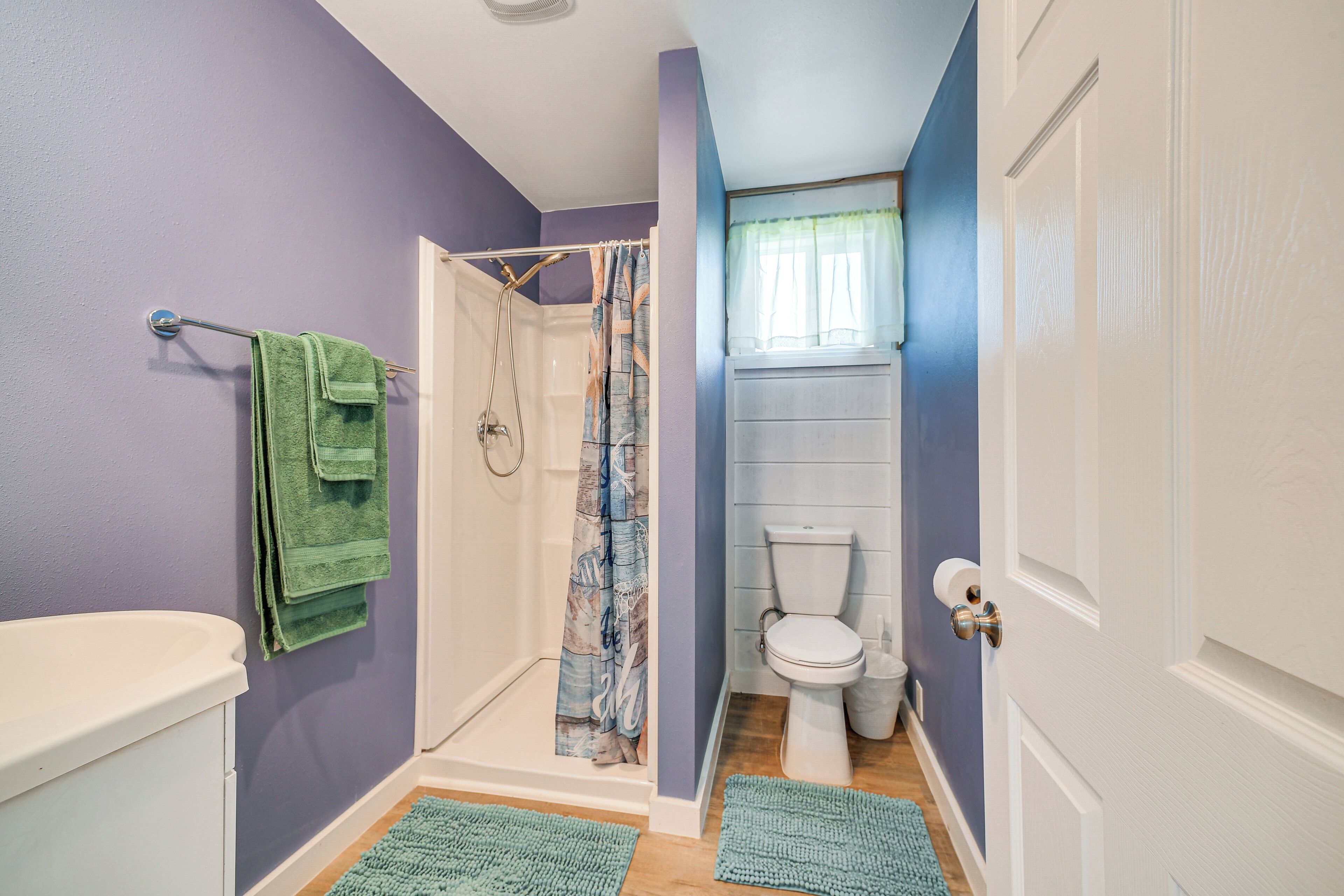 A bathroom with purple walls, a white toilet, and a shower with a curtain. There are green towels hanging on a rack and a white door.
