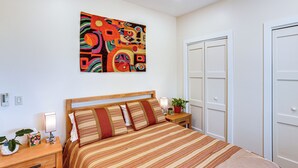 Une chambre à coucher bien rangée, avec un grand lit, une table de chevet avec une lampe, une plante en pot et un tableau abstrait aux couleurs vives accroché au mur.