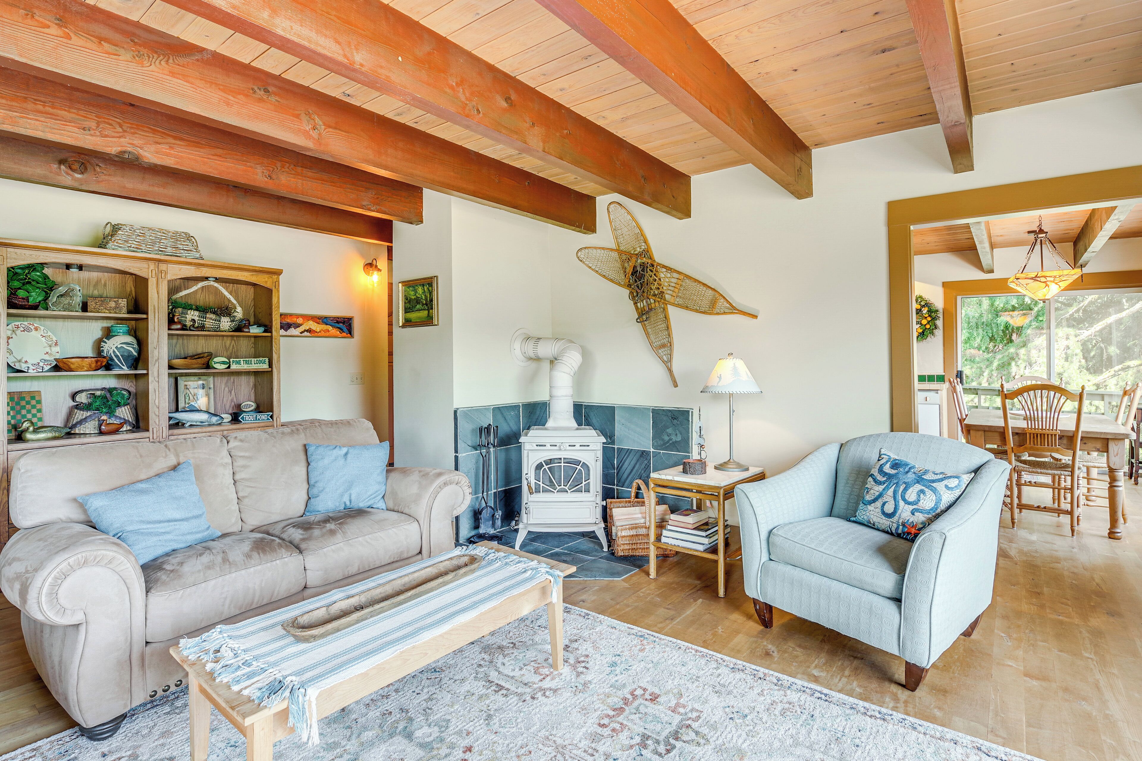 A cozy living room with a sofa, armchair, and a small table. A white stove is in the corner, and there's a large window with a view of greenery.