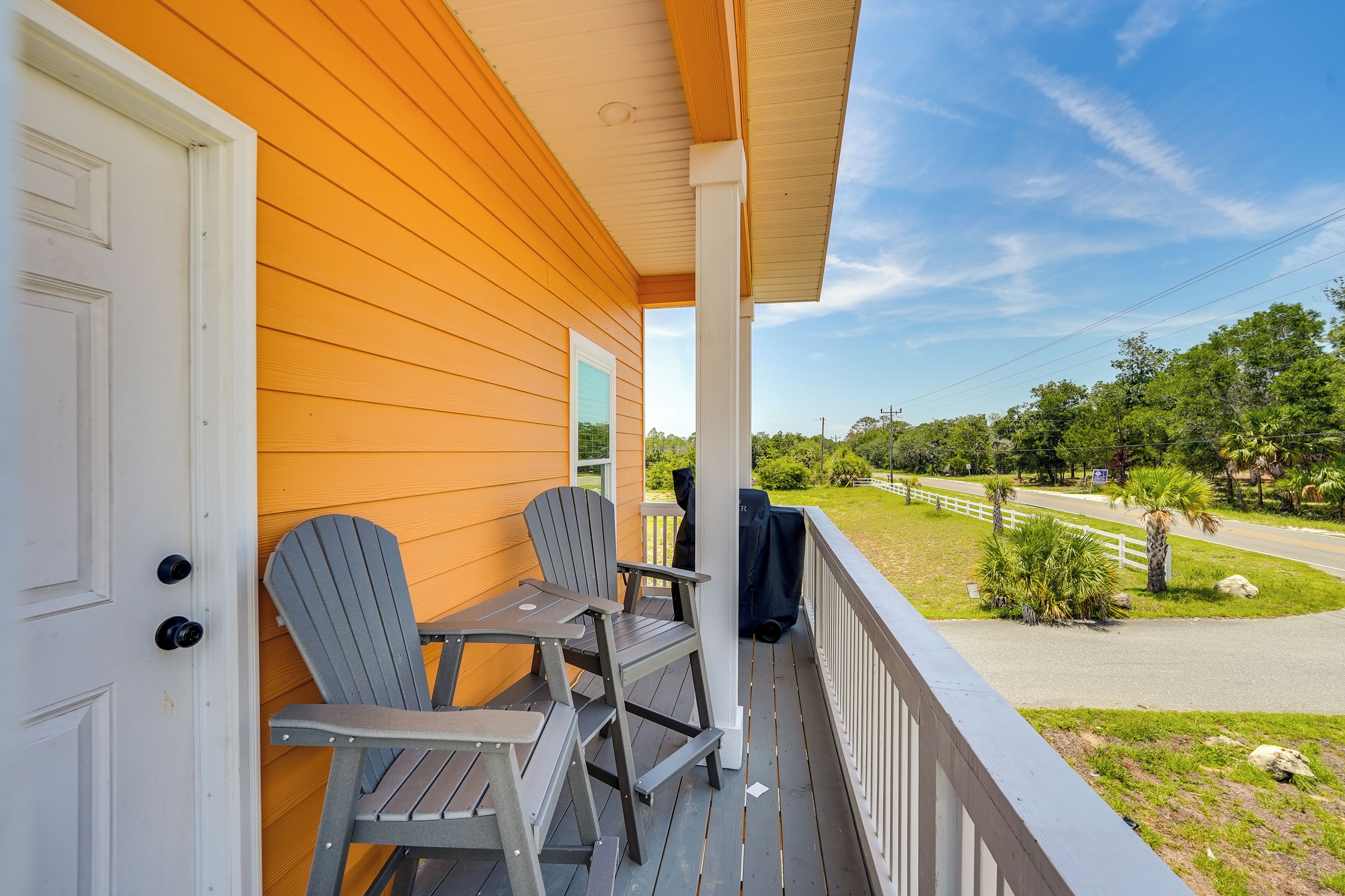 Eine Veranda mit zwei Adirondack-Stühlen und einem Tisch, mit Blick auf eine Straße, gesäumt von Grünflächen und einem weißen Zaun.